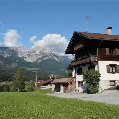 Ein idyllisches Haus in den Bergen mit grünen Wiesen und majestätischen Gipfeln im Hintergrund. Der Himmel ist klar und sonnig.