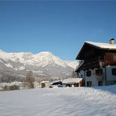 Eine malerische Winterlandschaft mit schneebedeckten Bergen im Hintergrund. Im Vordergrund steht ein traditionelles Holzhaus.