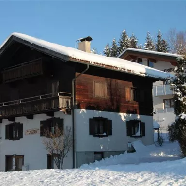 Ein traditionelles Chalet im Schnee, umgeben von Bäumen. Der Himmel ist klar und die Sonne scheint durch die winterliche Landschaft.