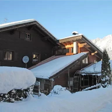 Ein traditionelles Chalet im Schnee mit einem klaren blauen Himmel. Im Hintergrund sind schneebedeckte Berge zu sehen.