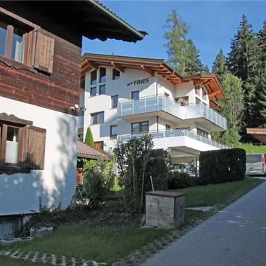 Ein modernes Gebäude mit einem Balkon, umgeben von Bäumen. Im Vordergrund steht ein traditionelles Holzhaus und eine gemütliche Straße.