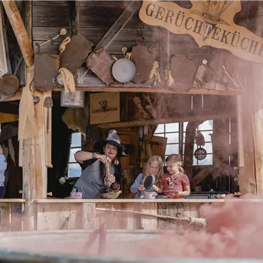 A rustic hut with the sign "Smell Kitchen" and several children watching curiously. A woman is preparing something as colorful vapors form around her.