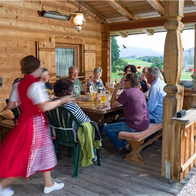A group of people is sitting at a wooden table on a porch, enjoying a meal together. In the background, green fields and flowers can be seen.