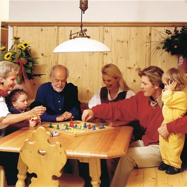 Eine Familie spielt gemeinsam ein Brettspiel am Tisch. Umgeben von Holzvertäfelung und Blumenstrauß, genießen sie die Zeit miteinander.