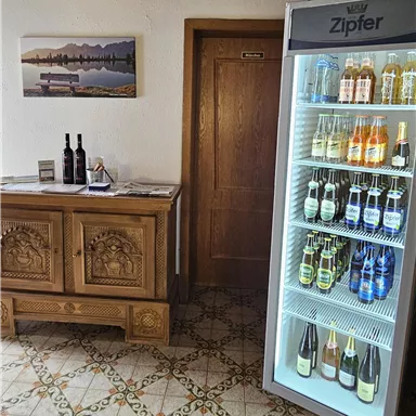 A cozy room with a wooden dresser and a refrigerator full of drinks. On the wall hangs a picture of mountains and lakes.