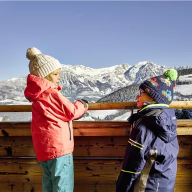 Two children are standing by a wooden barrier, looking at a snow-covered mountain landscape. They are wearing warm clothes and hats while the clear blue sky shines above them.