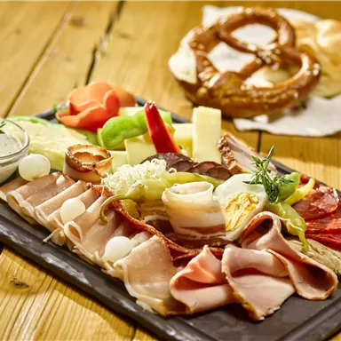 A varied plate with sausage, cheese, and fresh vegetables. In the background, a pretzel and a bread roll are visible.