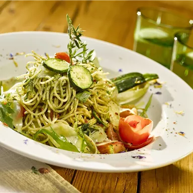 A delicious plate of pasta with fresh vegetables and herbs. In the background, there are two green drinks.