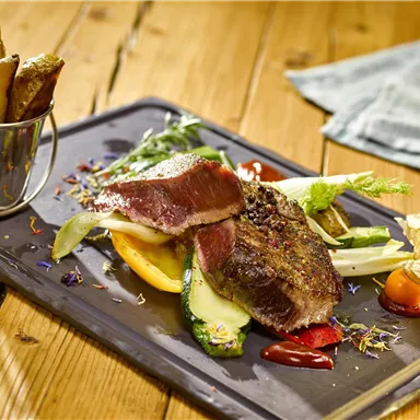 A plate with tender meat, colorful vegetables, and a side of baked potatoes. The dish is presented on a black tray and looks appetizing.