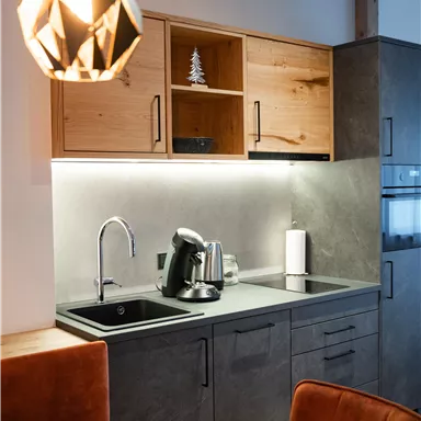 Eine moderne Küchenzeile mit Holzregalen und dunklen Oberflächen. Eine elegante Küchenlampe hängt über einem Tisch mit orangefarbenen Stühlen.