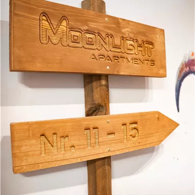 Ein Holzschild mit der Aufschrift „Moonlight Apartments“ und der Richtung zu den Nummern 11-15. Das Schild ist einfach gestaltet und verleiht einen rustikalen Eindruck.