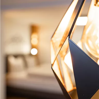 Eine moderne Lampe in geometrischer Form hängt im Vordergrund. Im Hintergrund ist ein helles, stilvolles Zimmer zu sehen.