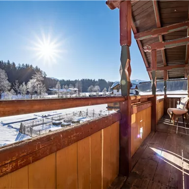 A balcony with a wooden railing that offers a view of a snowy landscape. The sun shines bright in the sky, creating a bright, inviting atmosphere.