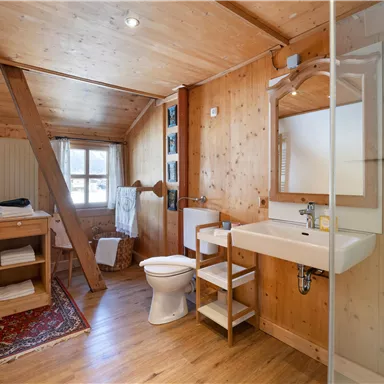 A cozy bathroom with wooden walls and a large window. It features a shower, a sink, and a toilet.