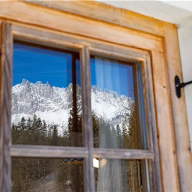 A window with a wooden frame that reflects the snow-covered mountains and the clear blue sky. Forests are also visible in the background.