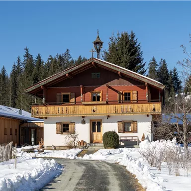 A cozy wooden house with a red facade and a balcony. It is located in a snow-covered landscape, surrounded by trees.