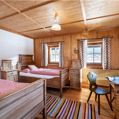 A cozy wooden bedroom with two beds, a table, and chairs. The walls are paneled with wood, and large windows let in plenty of light.