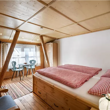 A cozy bedroom with wooden paneling and two single beds. In the background, there is a table and chairs as well as a window with curtains.