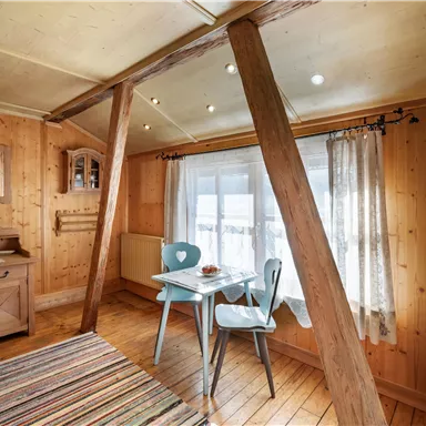 A cozy room with paneled wooden walls and floor. A small table with two chairs stands in front of a window that lets in a lot of daylight.