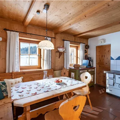 A cozy dining area in a wooden house with a long table and chairs. The walls are made of wood, and large windows let in plenty of light.