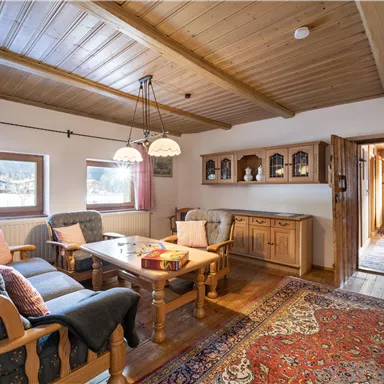 A cozy living room with wooden ceilings and a large table. The decor is rustic with comfortable armchairs and a colorful rug.