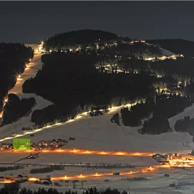 Eine verschneite Landschaft bei Nacht mit beleuchteten Skipisten und kleinen Häusern. Der Himmel ist dunkel und die Lichter der Ortschaft erhellen die Szene.