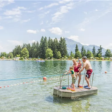 A small pier in the water with two adults and a child having fun. In the background, there are trees and mountains visible, in nice weather.