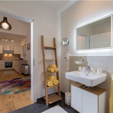Ein modernes Badezimmer mit einem stilvollen Waschbecken und einem beleuchteten Spiegel. Im Hintergrund sieht man eine offene Küche mit einem Teppichboden.