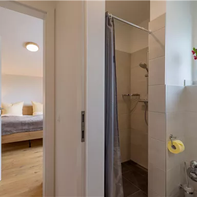 Ein modernes Badezimmer mit einer Dusche und einer kleinen Blume auf der Fensterbank. Im Hintergrund ist ein Schlafzimmer mit einem Bett zu sehen.