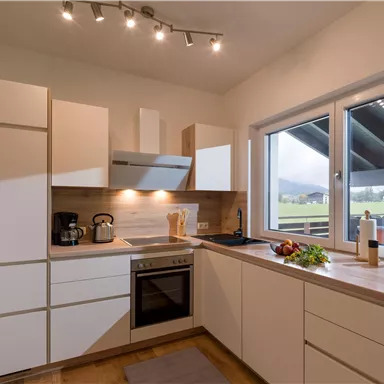Eine moderne Küche mit weißen Möbeln und einer großen Fensterfront. Der Raum ist hell und einladend, mit einem Blick auf die Landschaft außerhalb.