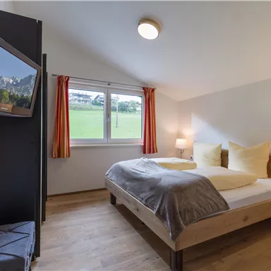 Ein gemütliches Schlafzimmer mit einem Doppelbett und großen Fenstern. Die Wände sind in neutralen Farben gehalten und es gibt einen Fernseher.