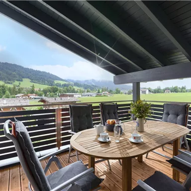Ein gemütlicher Balkon mit einem runden Holztisch und fünf Stühlen. Im Hintergrund sind grüne Wiesen und Berge zu sehen.