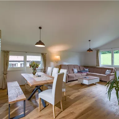 Ein modernes und helles Wohnzimmer mit einem großen Esstisch und einer gemütlichen Couch. Große Fenster sorgen für viel Tageslicht und bieten einen Blick ins Grüne.