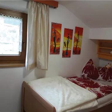 A cozy bedroom with a single bed and colorful wall paintings. The room has a window with a view outside and a friendly, bright atmosphere.