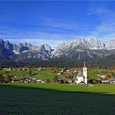 Eine malerische Landschaft mit hohen Bergen und einem kleinen Dorf. Im Vordergrund steht eine Kirche, umgeben von grünen Wiesen und traditionellen Häusern.