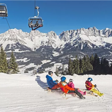 Eine Familie genießt einen sonnigen Tag im Schnee. Im Hintergrund sind majestätische Berge und ein Skilift zu sehen.