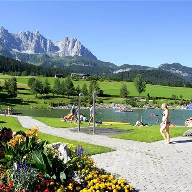 Ein schöner See umgeben von grünen Wiesen und Bergen. Menschen genießen die Sonne und entspannen auf Liegen.
