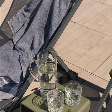 A deck chair with a blue towel and a small table. On it are a pitcher of water and several glasses.