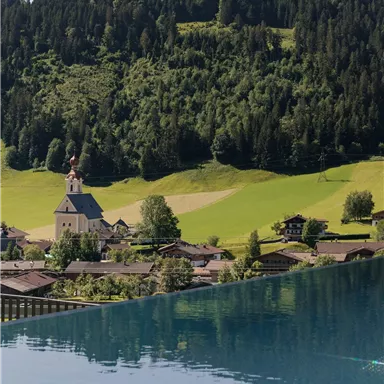 A picturesque village with a church, surrounded by green meadows and forests. In the foreground, the water of a pool reflects.