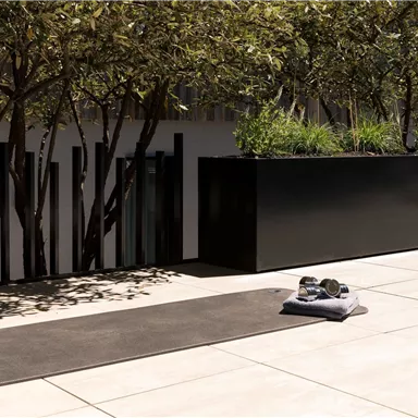 A modern, bright outdoor area with a yoga mat on the floor. In the background, green plants and a black planter box can be seen.