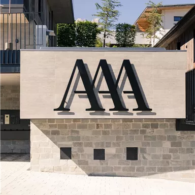 A modern building with a striking logo sign. The facade is made of light stone and is surrounded by green plants.