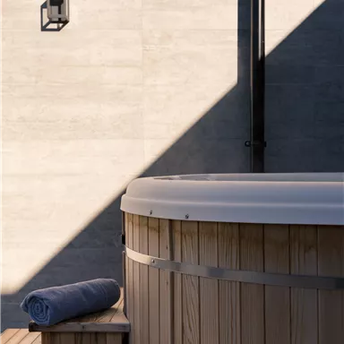 A wooden tub stands on a wooden deck, surrounded by a simple wall. A towel lies beside it, and soft light falls on the scene.