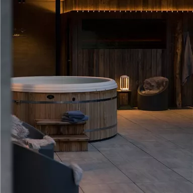 A modern outdoor area with a wooden hot tub and a cozy seating arrangement. Soft lighting creates a relaxed atmosphere.