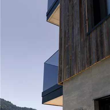 A modern building with wooden elements and glass balconies. In the background, green hills and a blue sky can be seen.