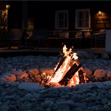 A cozy bonfire in a stone fire pit. The background is dimly lit with silver and wooden furnishings.