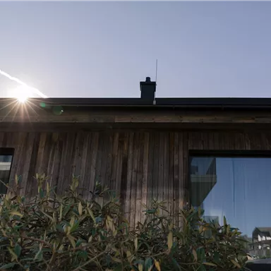 A modern wooden house under a clear sky. The sun shines out from behind the roof.