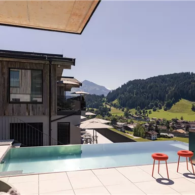 A modern terrace with an infinity pool and two red bar stools. In the background, a picturesque mountain landscape with green meadows stretches out.