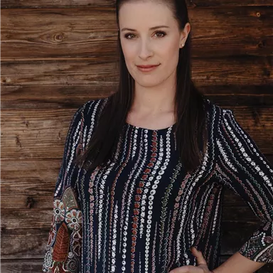 A woman is standing in front of a wooden wall and wearing a patterned top. She has long hair and is looking directly at the camera.
