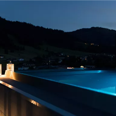 A modernly designed infinity pool at night, surrounded by gentle hills. Warm lights create an inviting atmosphere.