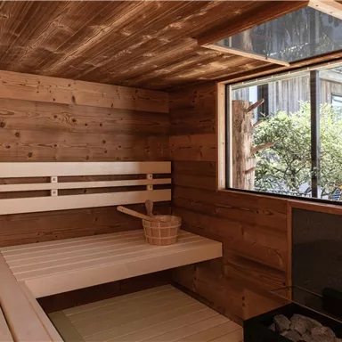 A cozy wooden sauna with benches and a window view into the greenery. The room contains a bucket and stones for the sauna.